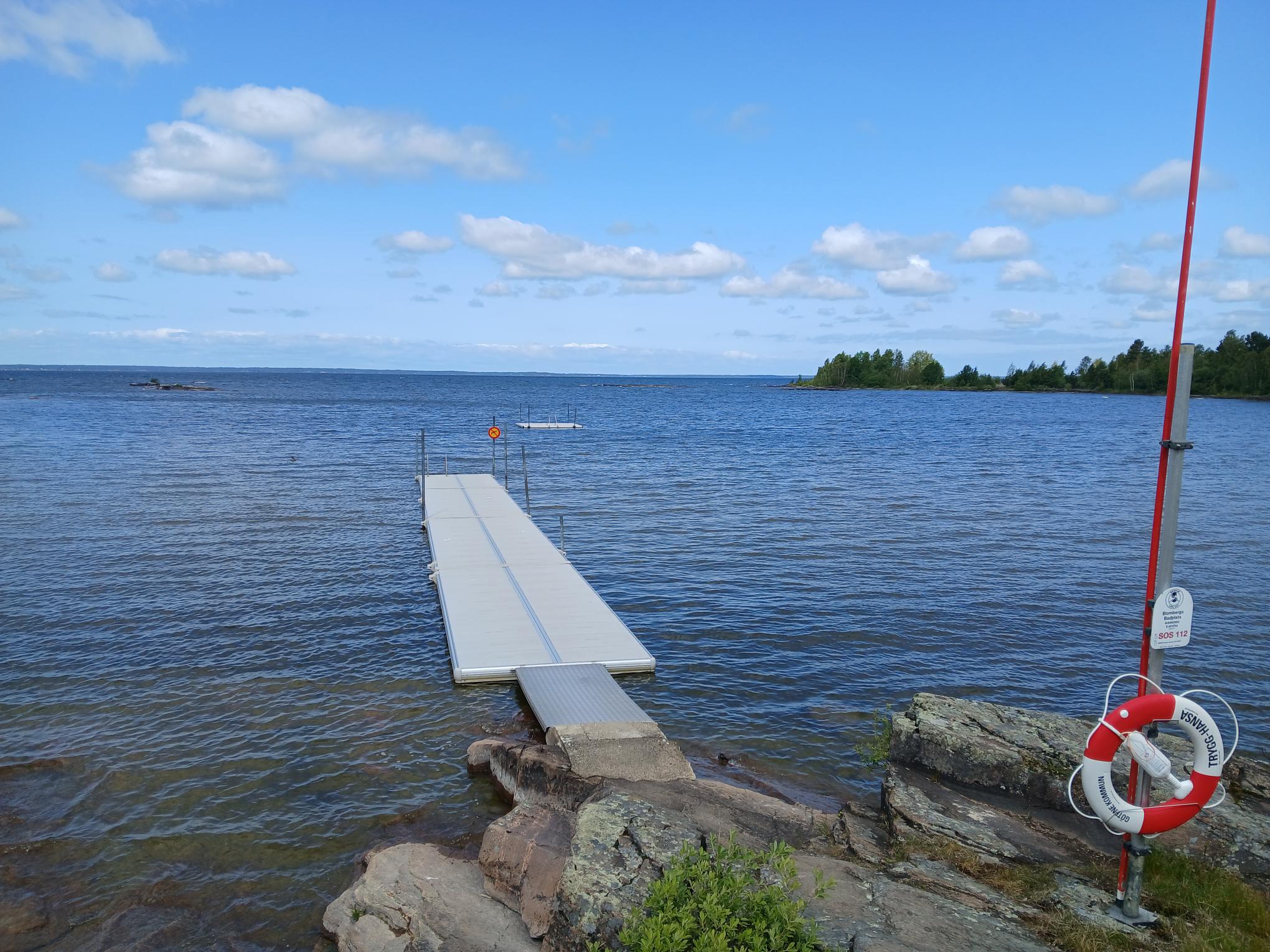 Blombergs badplats. En ljusgrå avlång flytbrygga som ligger i vattnetfastankrad i ett berg med en metallstege ner till vattnet. Till höger hänger en röd-vit räddningsflytring att använda.