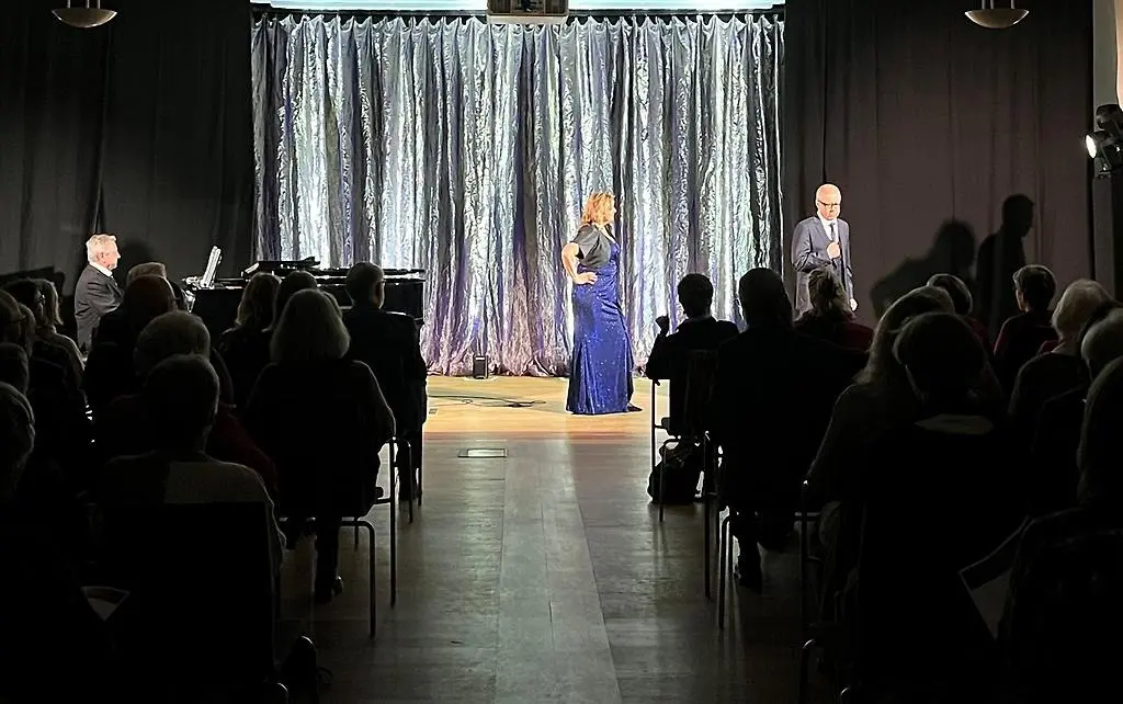 Publik som sitter och tittar på en kvinna med ljust hår och lila långklänning som står och sjunger tillsammans med en kostymklädd man vid sin sida. På scenen finns också en man som spelar på en svart flygel. Scenen är upplyst och ett glänsande, silvrigt draperi hänger i bakgrunden.