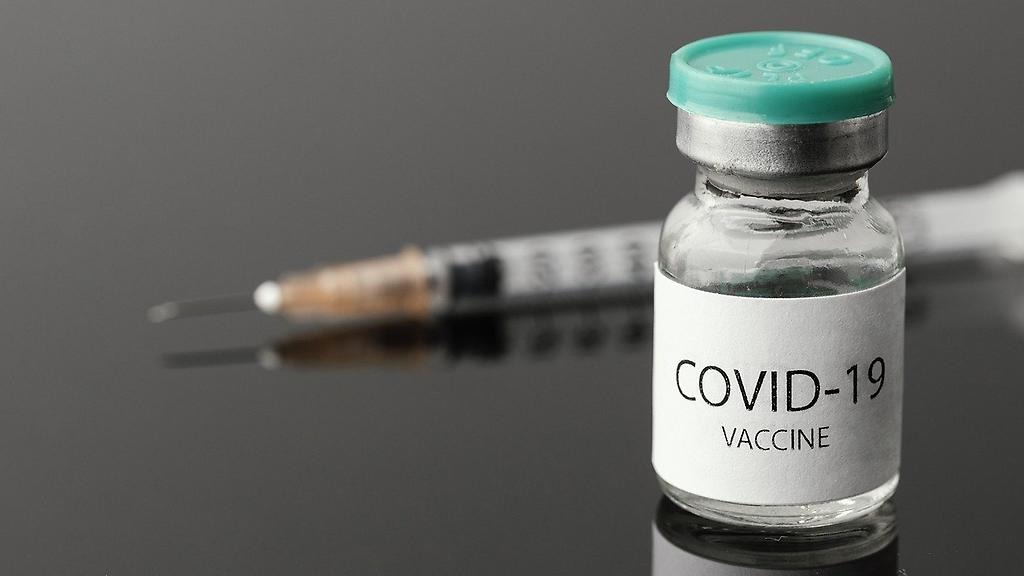 En glasflaska med texten COVID-19 VACCINE på. I bakgrunden en spruta.