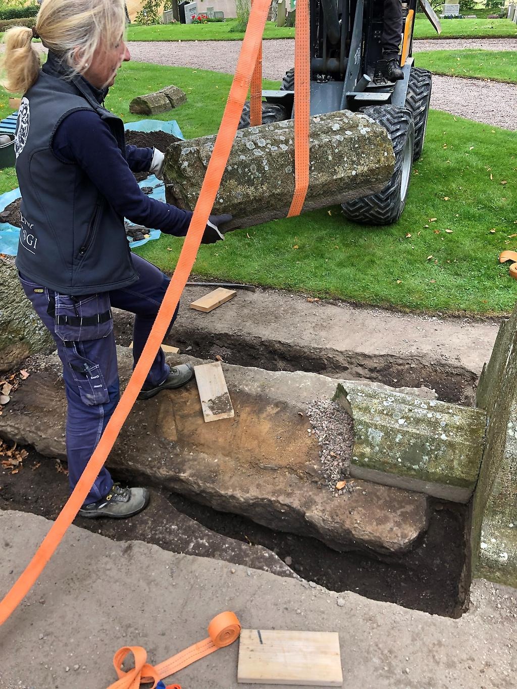Kvinna står i en utgrävning och en traktor lyfter upp en stor stenbit.