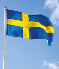 Svensk flagga i toppen på en flaggstång omgiven av träd.
