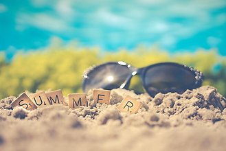 Svarta solglasögon liggandes på en beige strand och små träbrickor framför med texten SUMMER.