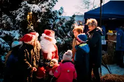 Jultomte framför snöklädd gran med förväntansfulla barn omkring sig.