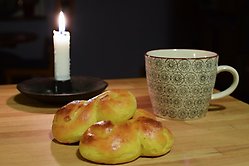 Ett träbord med ett brinnande ljus, 2 stycken lussekatter och en kaffemugg. 